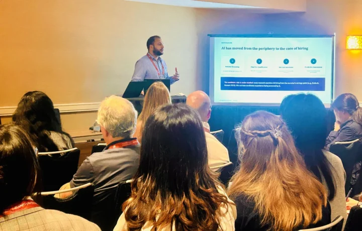 Presenting to a seated audience at the Academy of Management Annual Meeting 2026, beside a slide reading 'AI has moved from the periphery to the core of hiring'